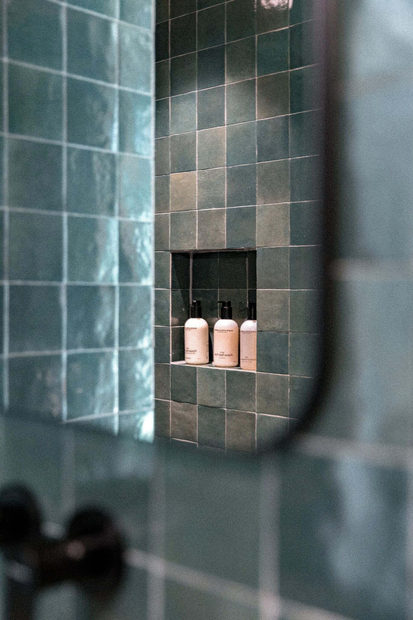 A bathroom with a tiled wall and a shelf containing three bottles.