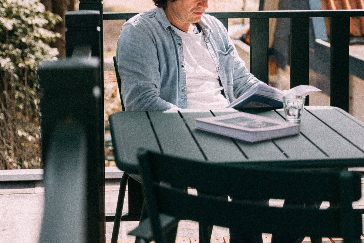 Eine Person sitzt an einem Tisch mit einem Buch und einem Glas Wasser, draußen vor Bäumen und einem Gebäude im Hintergrund.