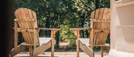 Dos sillas Adirondack de madera con una pequeña mesa entre ellas, ubicadas en un patio con vistas a una zona boscosa.