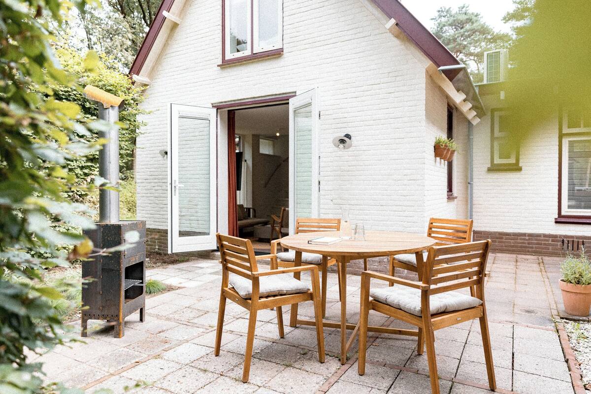 Eine Terrasse mit einem runden Holztisch und Stühlen, einem Grill und einem weißen Haus mit rotem Dach.