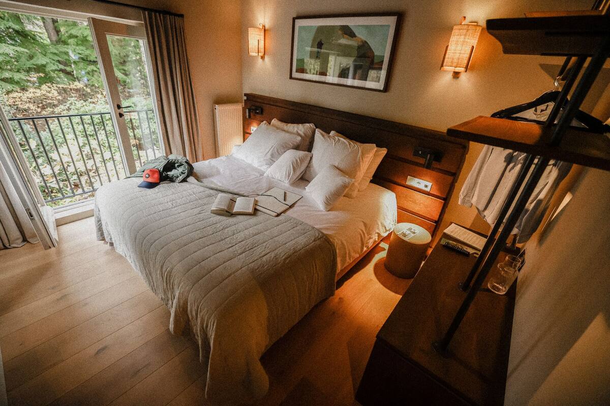 Ein Hotelzimmer mit einem großen Bett, einem Nachttisch, einer Lampe und Blick ins Freie.