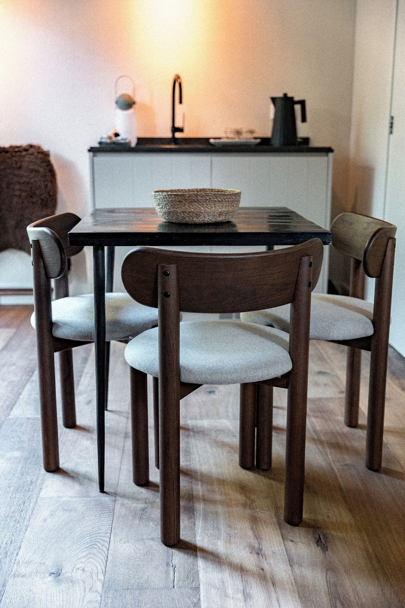 Un comedor con una mesa redonda, cuatro sillas, una cesta tejida, una tetera y una cafetera sobre un mueble blanco.