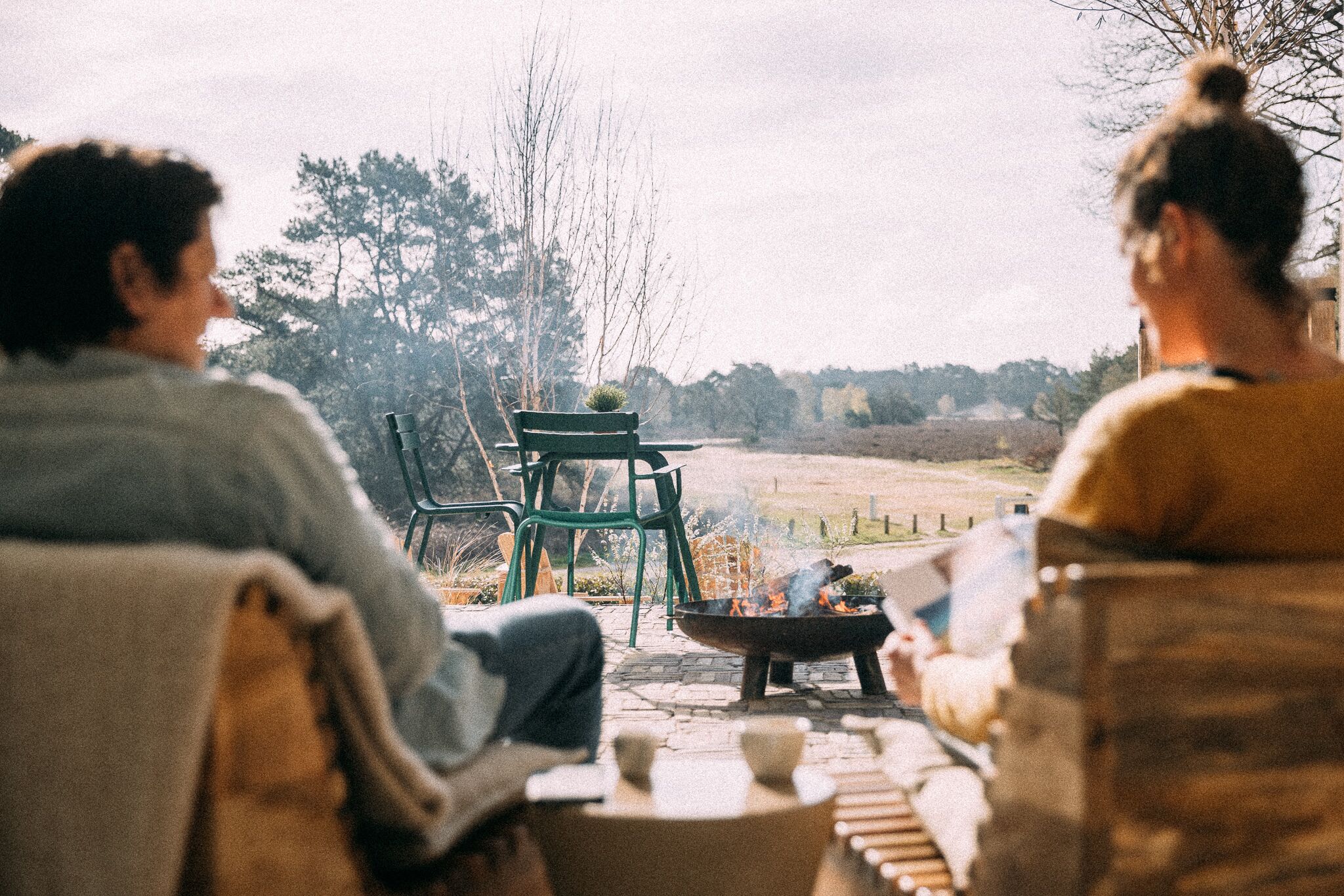 Two people sitting on outdoor furniture facing a fire pit with a green chair and a small table nearby.