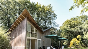 A wooden cabin with a large glass door, outdoor seating, and a green umbrella. - De Wever Lodge (Otterlo)