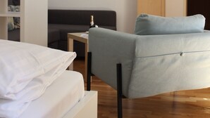 Ein modernes Hotelzimmer mit einem Bett, einem grauen Sofa, einem Holzkasten und einem kleinen Tisch mit einer Flasche.