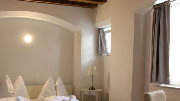 Ein Hotelzimmer mit Holzboden, einem Bett mit weißen Leinentüchern, einem Nachttisch mit Lampe und einem Fenster mit Vorhängen.