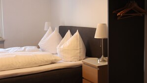 Una cama bien hecha con sábanas y almohadas blancas, una mesita de noche con una lámpara y un armario de madera con perchas.