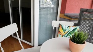 Una silla blanca y una mesa con una planta en maceta en un balcón con puerta de cristal corredera y una silla de metal.