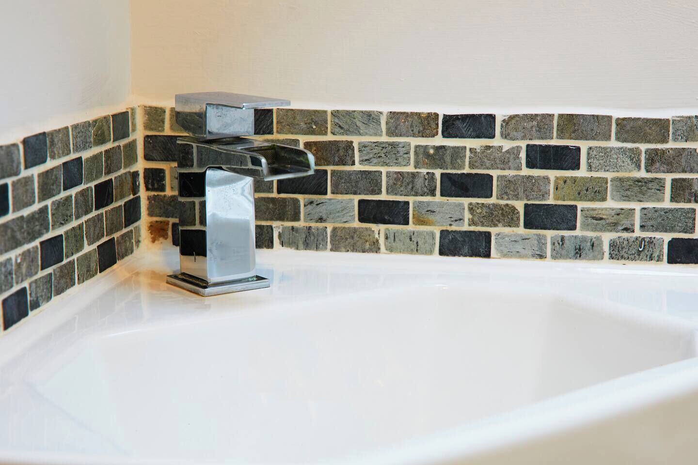 A corner of a bathroom with a white bathtub, a chrome faucet, and a mosaic tile wall.