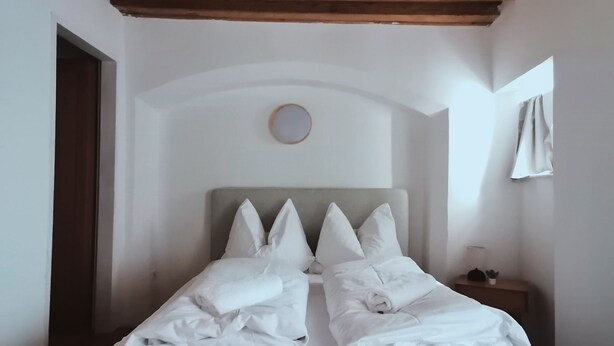 A bedroom with a wooden ceiling, a bed with white bedding, and a small bedside table with a lamp.