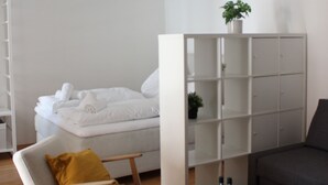 A modern hotel room with a bed, a seating area with a chair and a table, a bookshelf, and a potted plant.