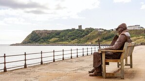 A statue of a seated figure on a bench by a waterfront with a hill and buildings in the background. - This apartment is a 2 bedroom(s), 2 bathrooms, located in Scarborough, North Yorkshire. (Scarborough)