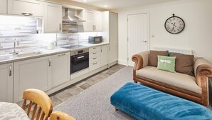 A modern kitchen with white cabinets, a microwave, and a built-in oven. There is a dining area with a table and chairs, and a blue ottoman. A clock is on the wall. - This apartment is a 2 bedroom(s), 2 bathrooms, located in Scarborough, North Yorkshire. (Scarborough)