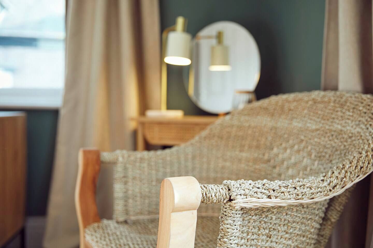A wicker chair with a wooden armrest in a room with a mirror and a lamp on a side table.