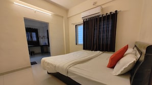 A bedroom with a bed, a window with curtains, an air conditioning unit, and a hallway leading to another room. - Vaishnav Inn (Pune)