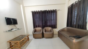 A living room with a sofa, two armchairs, a glass-top coffee table, a flat-screen TV, and a curtain. - Vaishnav Inn (Pune)