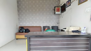 A reception desk with a dark wood finish and horizontal lines, a brown sofa with a black bag and a green pillow, and a wall-mounted TV. - Vaishnav Inn (Pune)