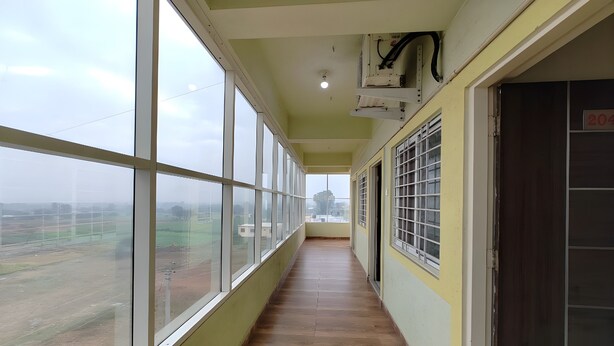 A corridor with large windows on one side, a wooden floor, and a door with a number sign on the other. - Vaishnav Inn (Pune)