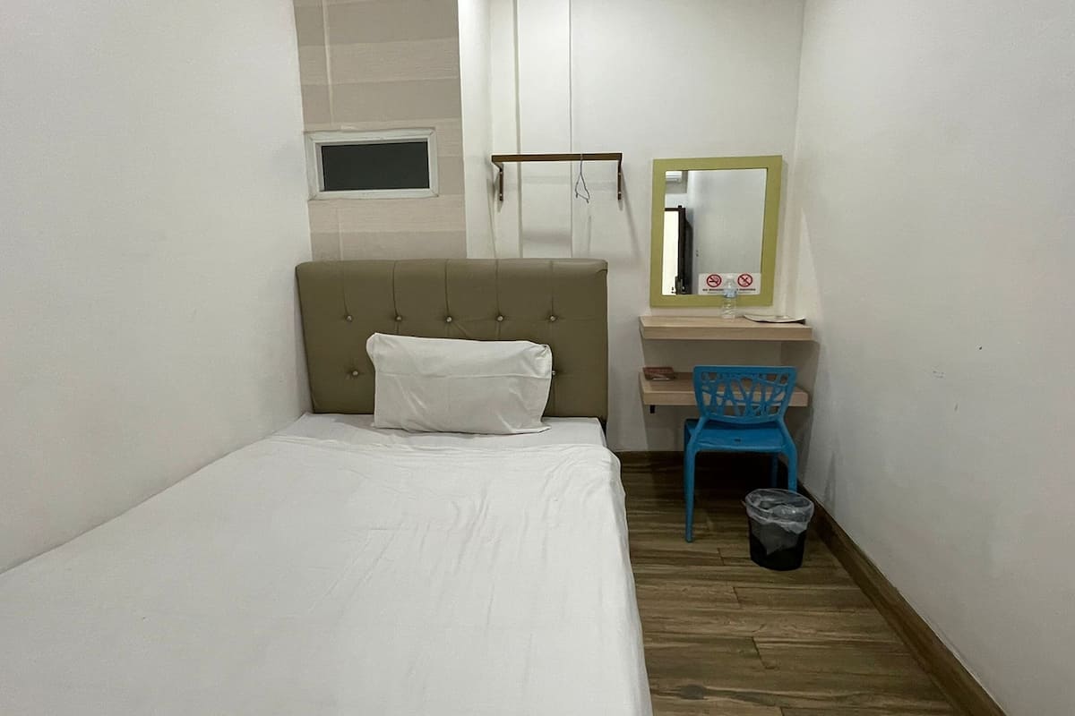A small, narrow room with a single bed, a blue chair, a small desk with a mirror, and a window.