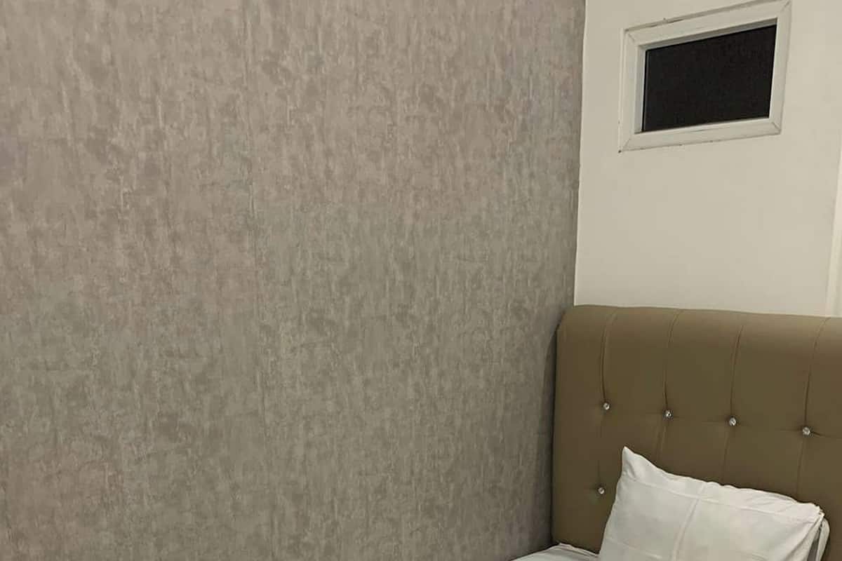 A single bed with a beige headboard and a white pillow in a room with a textured wall and a small window.
