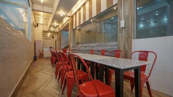 A dining area with a long table and red chairs, a wooden herringbone floor, and large windows with a view of the outside.