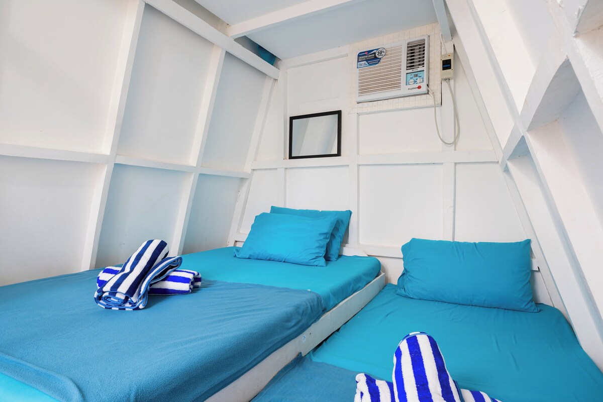 Une chambre avec deux lits, chacun doté de draps bleus et de serviettes à rayures. Il y a une fenêtre avec des stores et un climatiseur fixé au mur.
