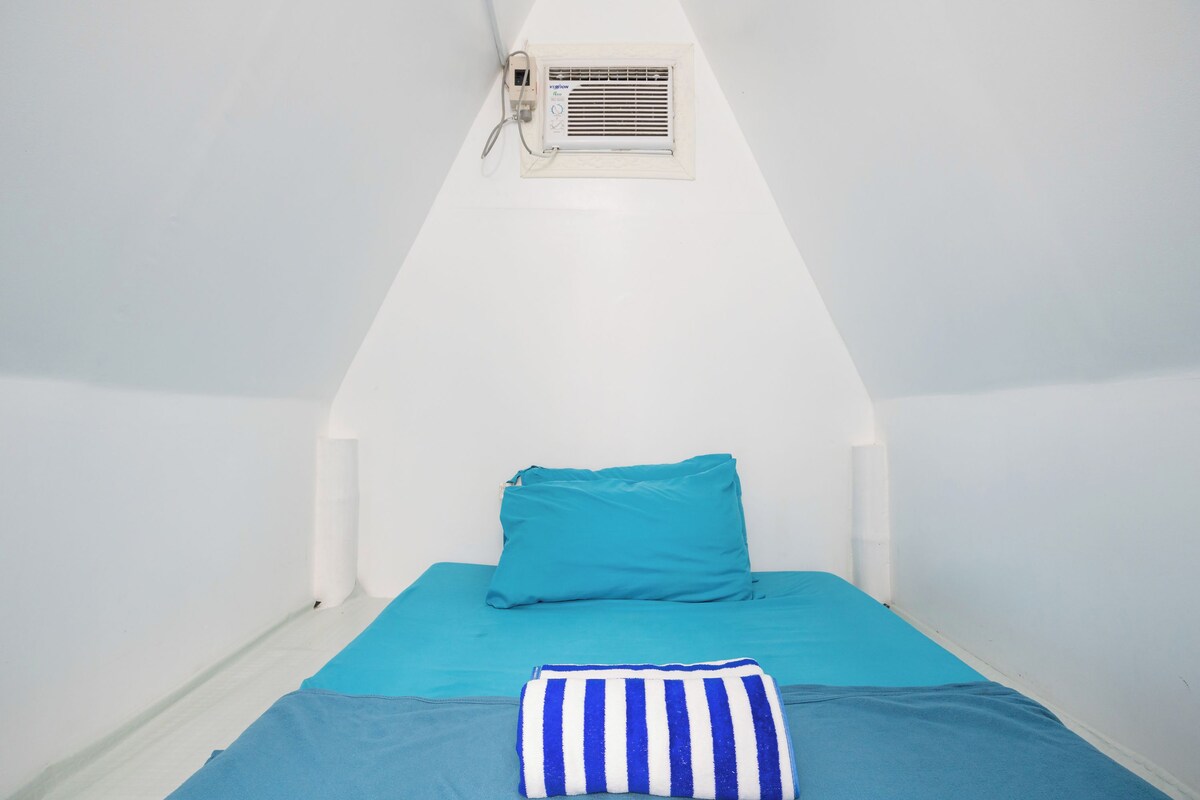 Een kleine eenpersoonskamer met een schuine kap, een raam met een airconditioning en een smaragdgroene spreidekbed.