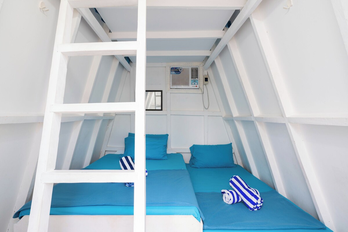 Een kamer met twee bedden, elk voorzien van blauw beddengoed en gestreepte kussens. Er is een witte ladder die naar een raam leidt, waar een airconditioningunit boven hangt.