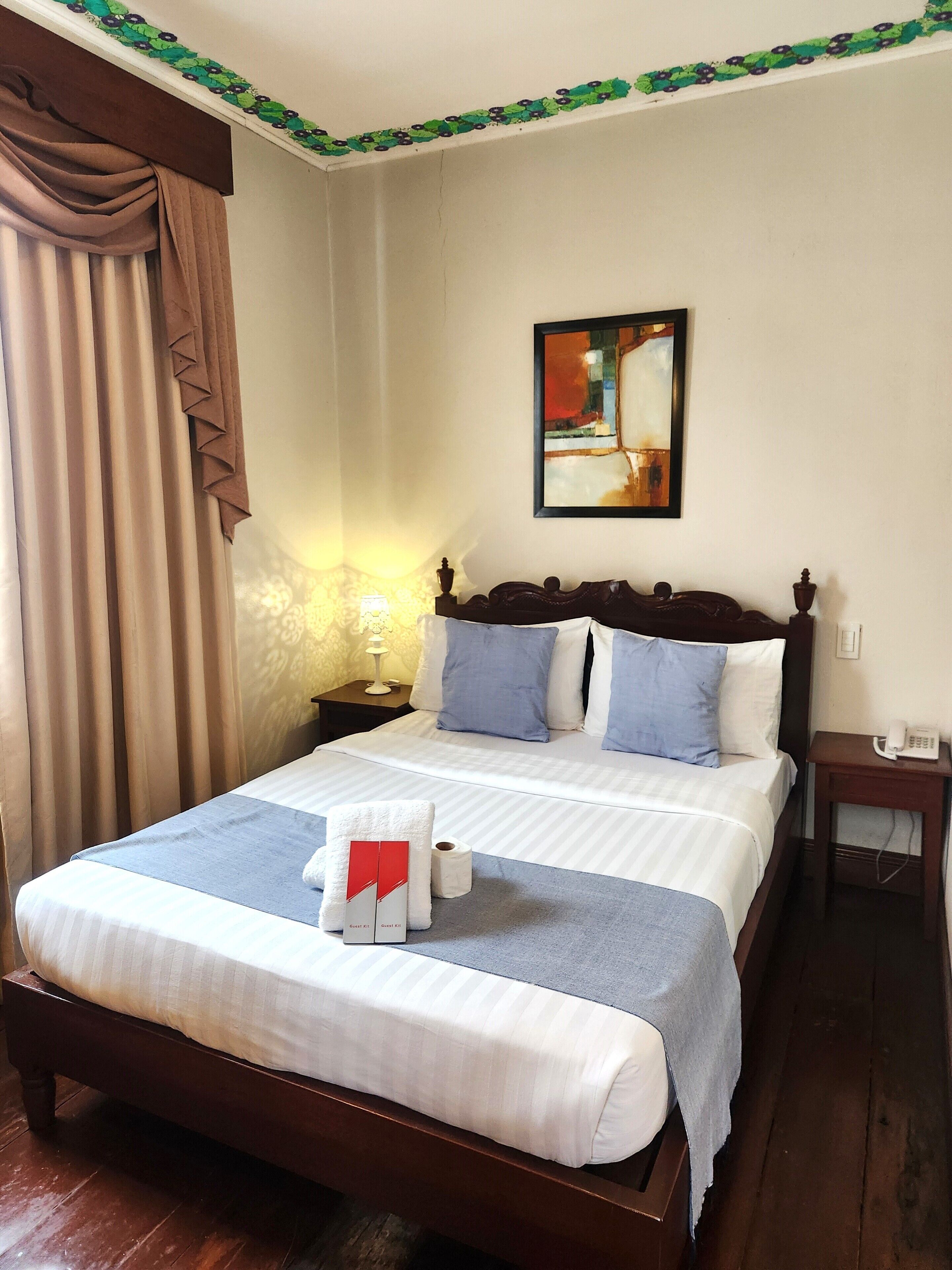 A neatly made bed with a blue and white striped bedspread, a white towel, and a red and white guest kit. There is a painting on the wall above the bed, a nightstand with a lamp, and a wooden floor.