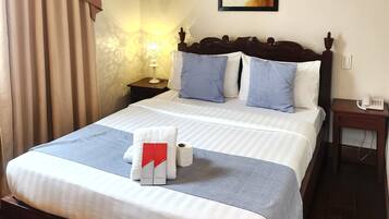 A neatly made bed with a blue and white striped bedspread, a white towel, and a red and white guest kit. There is a painting on the wall above the bed, a nightstand with a lamp, and a wooden floor.