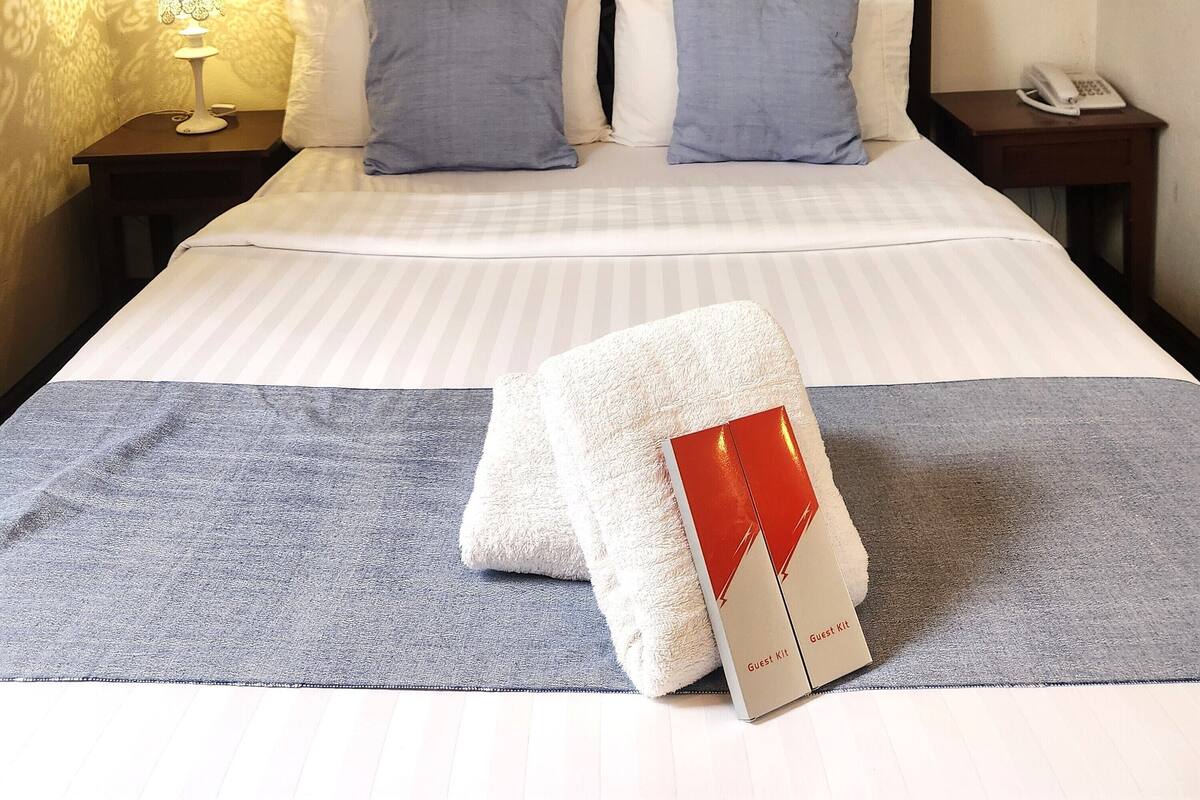 Una cama bien hecha con una colcha a rayas, dos almohadas y una toalla doblada sobre una alfombra gris. Un cuadro abstracto enmarco cuelga sobre la cama.