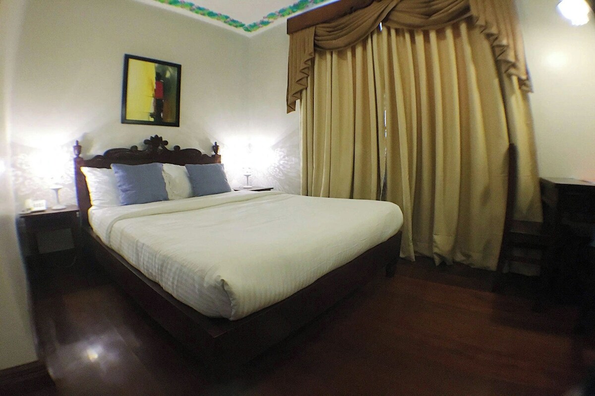 A neatly made bed with a wooden headboard, a bedside table with a lamp, and a large window with beige curtains.