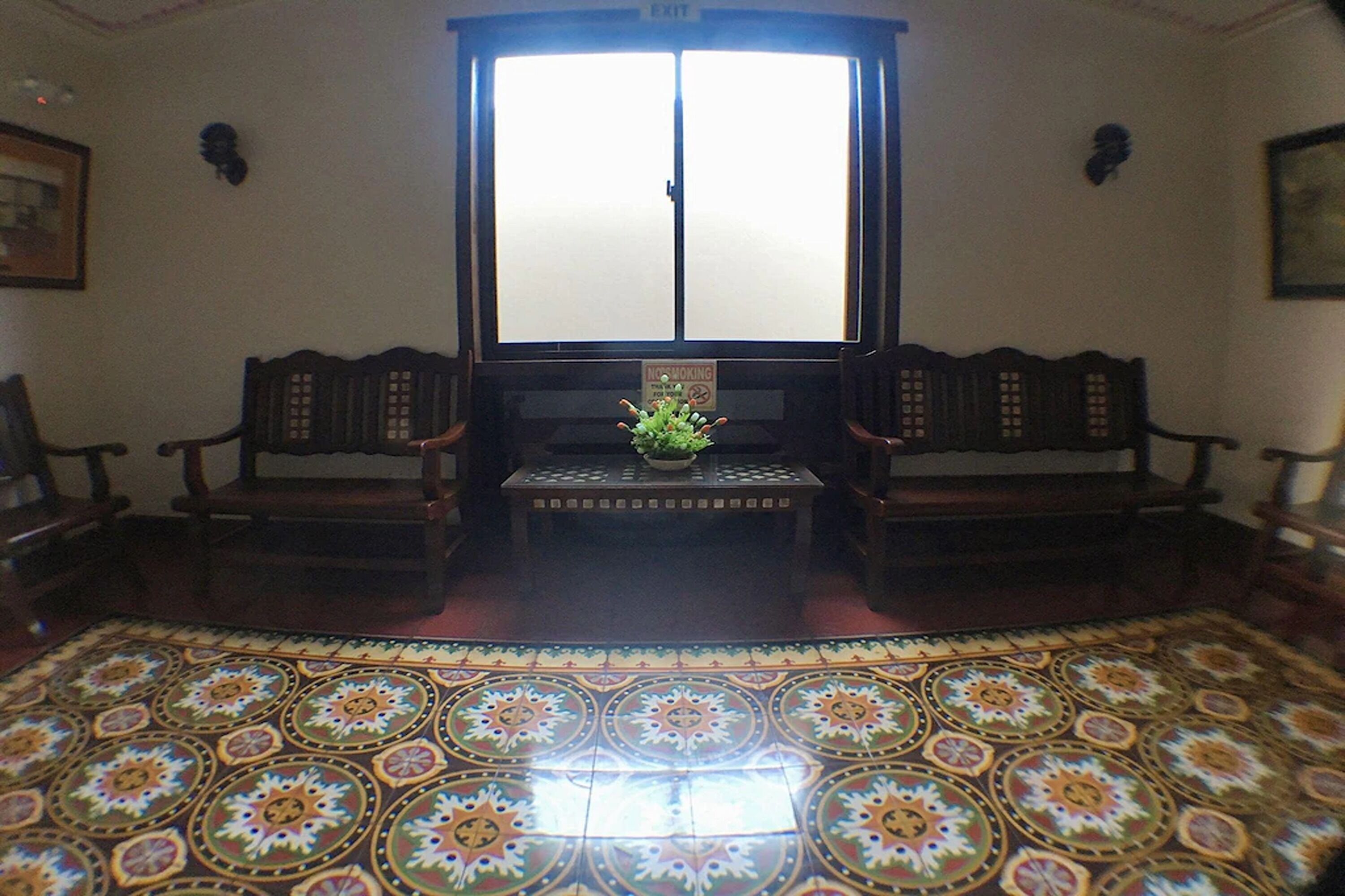 A room with a patterned floor, two wooden chairs, a small table with a plant, and a window with a 'No Smoking' sign.