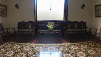 A room with a patterned floor, two wooden chairs, a small table with a plant, and a window with a 'No Smoking' sign.