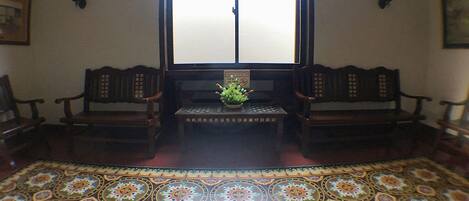 A room with a patterned floor, two wooden chairs, a small table with a plant, and a window with a 'No Smoking' sign.