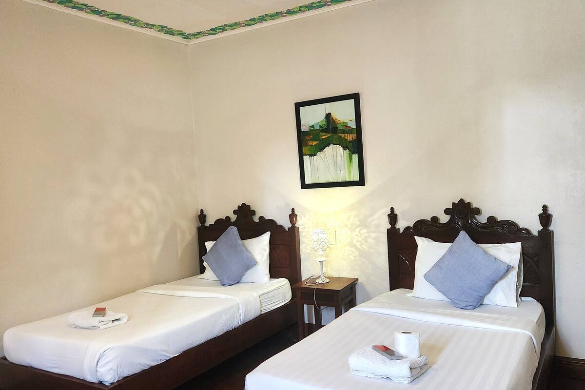 Habitación de hotel con dos camas individuales, cabeceros de madera, ropa de cama blanca, un cuadro en la pared y una mesita de noche con lámpara.