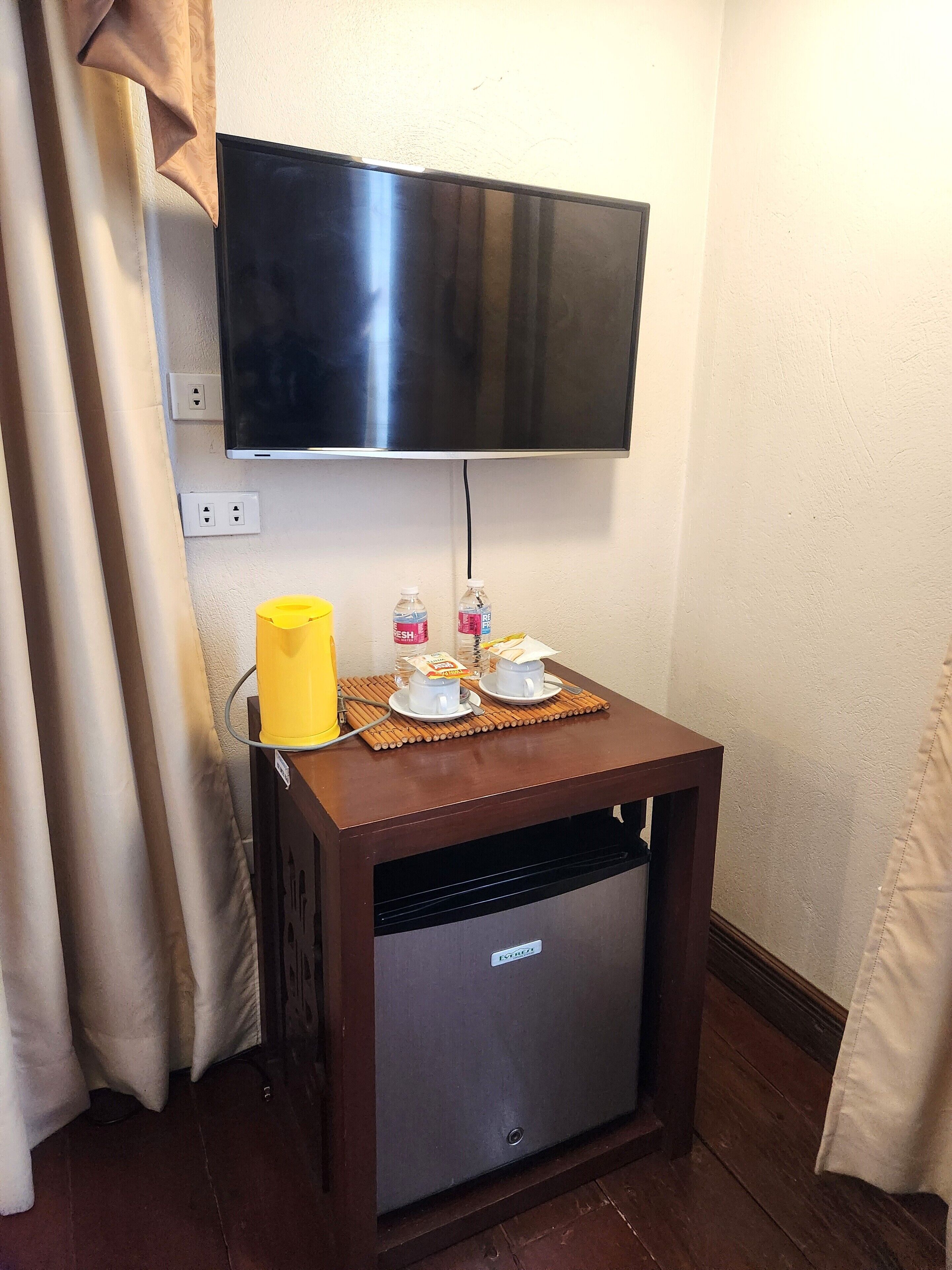 A small kitchenette with a TV mounted above it, a yellow water dispenser, and two white cups on a wooden tray.