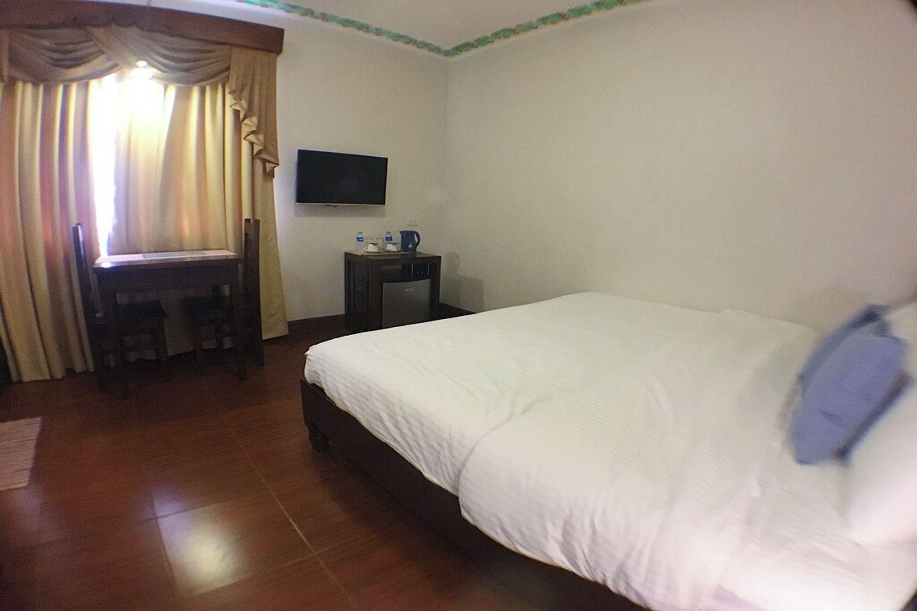 Habitación de hotel con cama, escritorio con silla, televisor y una mesita con jarrón y vasos.
