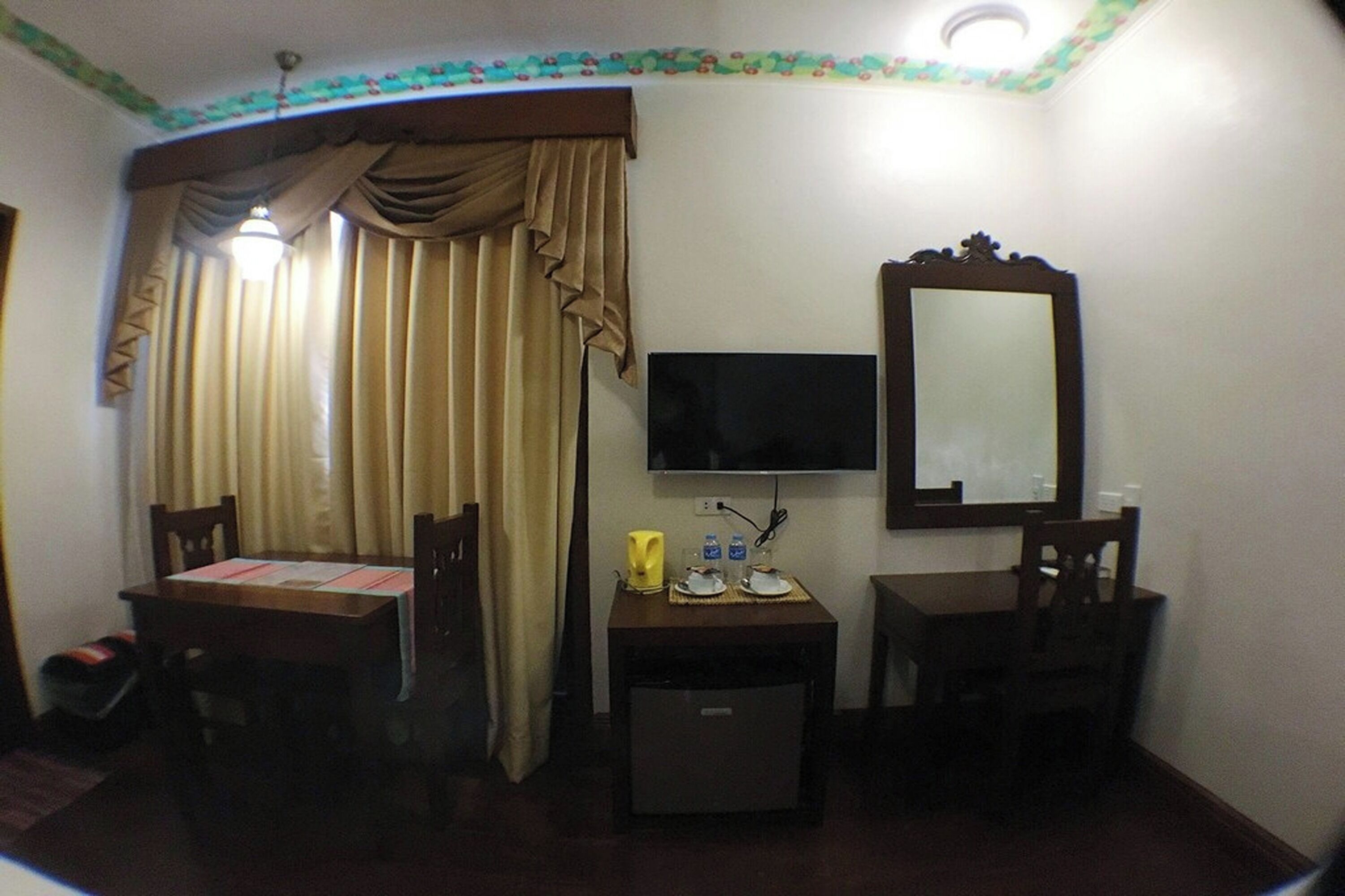 A room with a TV mounted on the wall, a small table with a mirror, and a window with curtains.