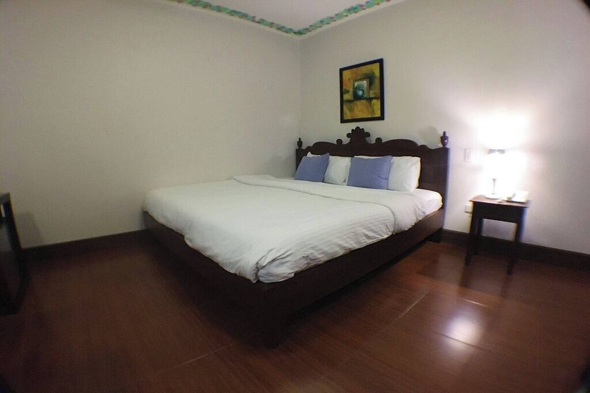 Un dormitorio con cama de madera, ropa de cama blanca, una mesita de noche con lámpara y un cuadro en la pared.