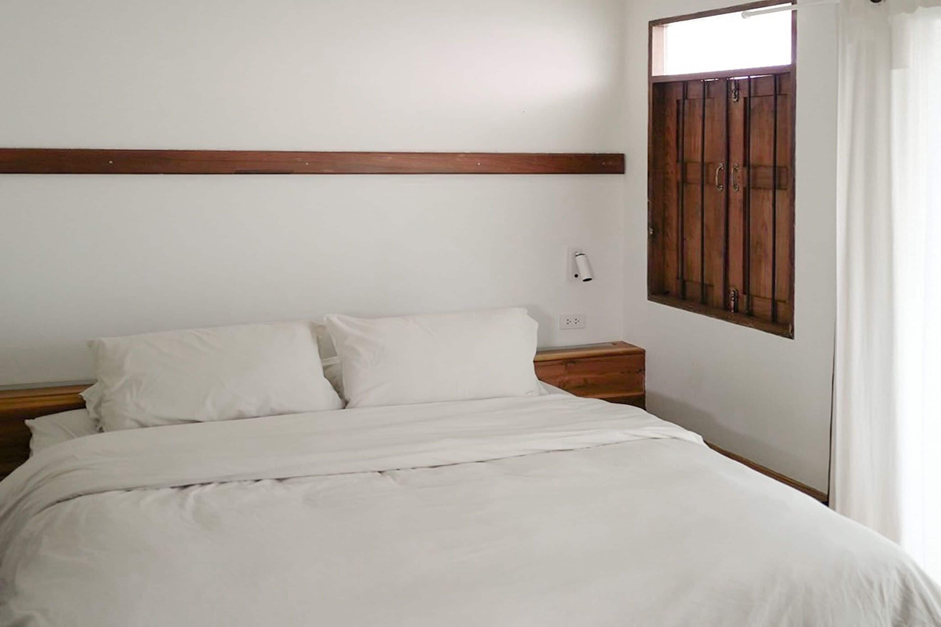 Una camera da letto con testiera in legno, un letto singolo con biancheria bianca, un comodino e una finestra con tende.