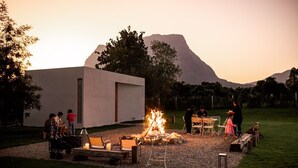 Un espacio al aire libre con fogón, rodeado de sillas y mesas, con una montaña y un edificio moderno de fondo.