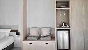Habitación de hotel pequeña y funcional con cama, un rincón de descanso con cojines, un mini refrigerador y una estantería con vasos y hervidor.