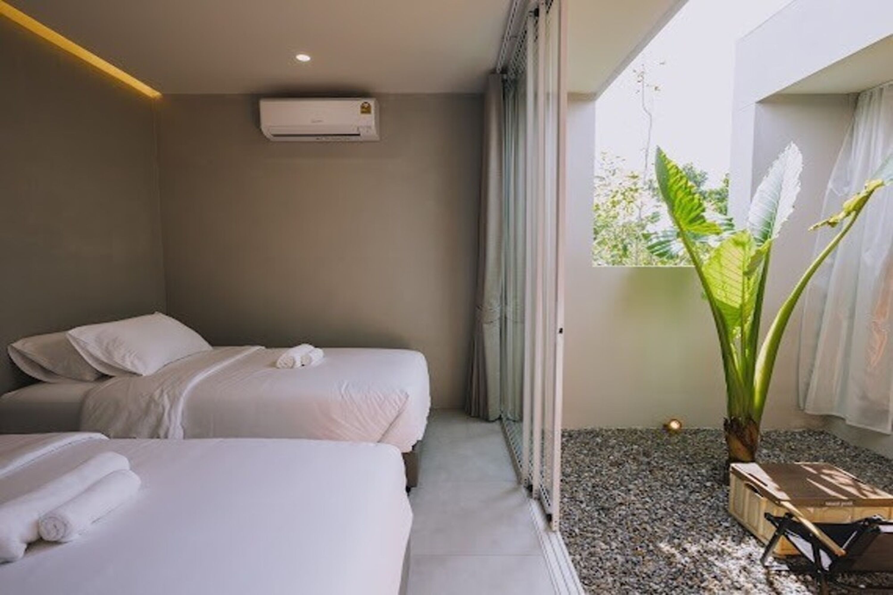 Habitación de hotel con dos camas, un ventanal con cortinas, un equipo de aire acondicionado y una planta en maceta.