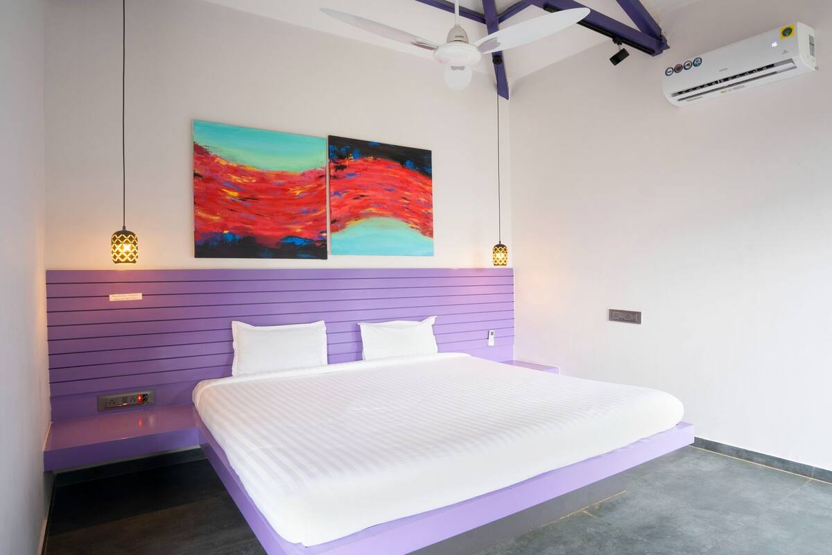 A modern bedroom with a purple headboard, a large bed, two wall-mounted paintings, a ceiling fan, and an air conditioning unit.