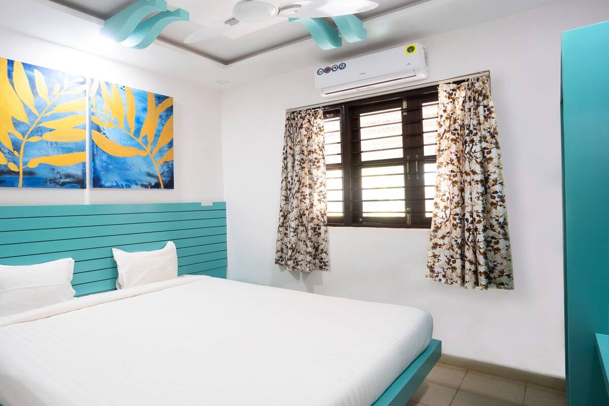 A bedroom with a bed, teal headboard, white bedding, two wall-mounted paintings, a window with floral curtains, and an air conditioning unit.