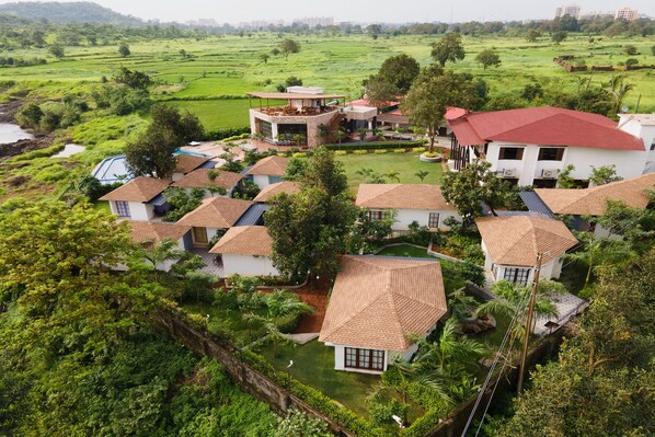 A residential area with multiple houses, some with red roofs and others with brown tiles, surrounded by lush greenery and fields. - Nirva The Cottage Resort, Kondeshwar Road Badlapur (Ambernath)