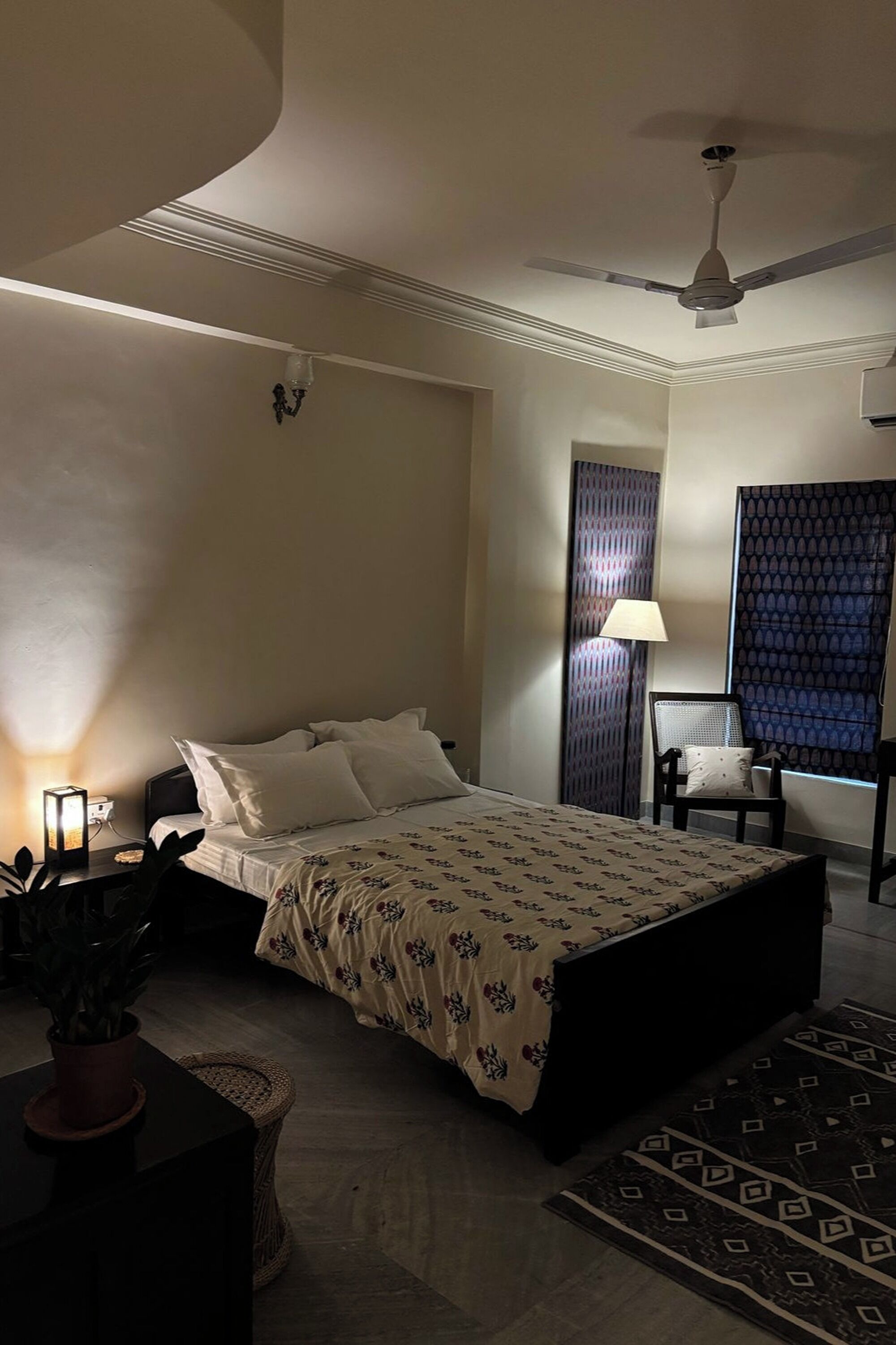 A bedroom with a bed, bedside lamp, chair, and a window with patterned curtains.