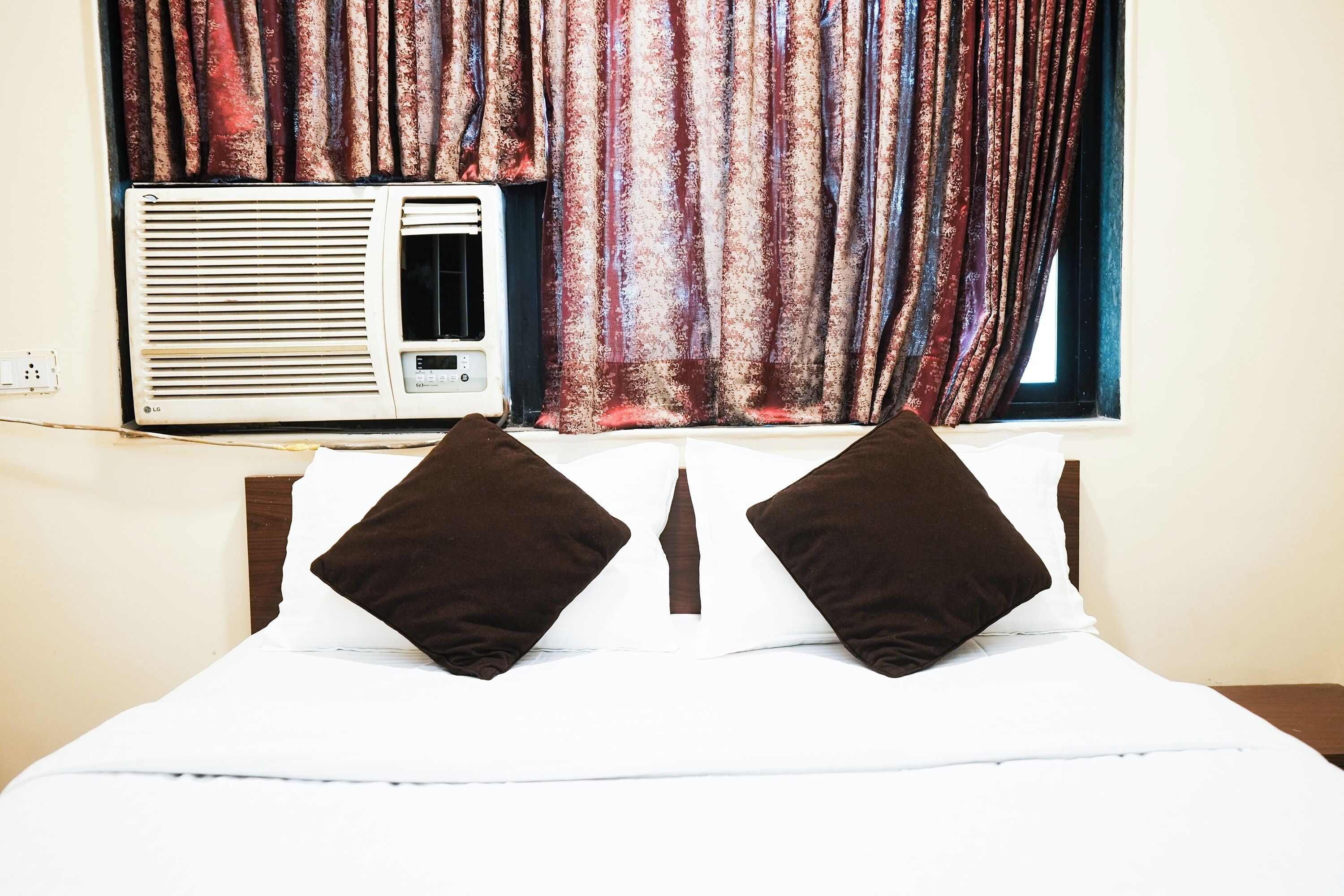 Ein Hotelzimmer mit einem Bett, zwei Kissen, einem Fenster mit Vorhängen und einer Klimaanlage.