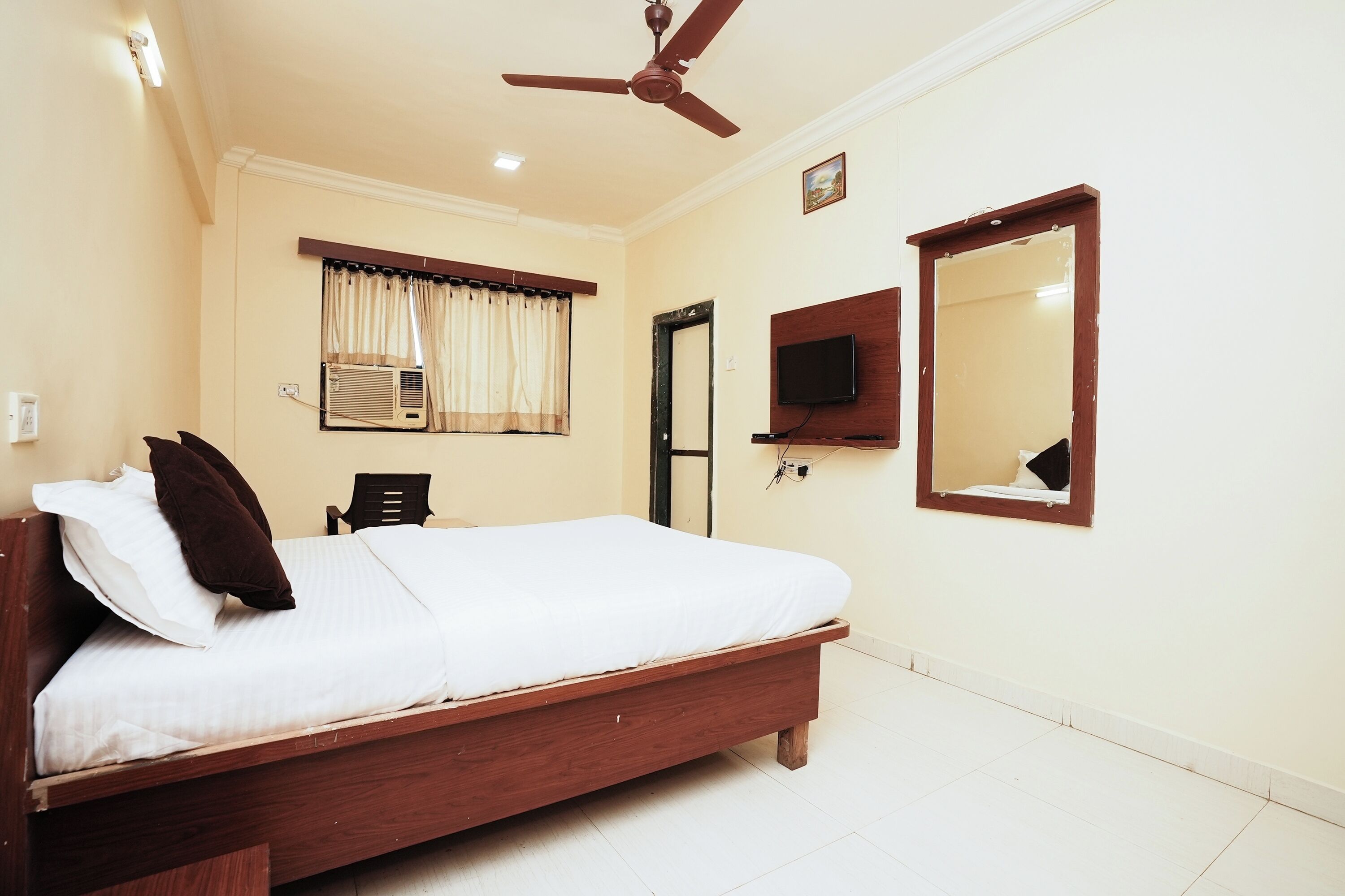 A bedroom with a wooden bed, a ceiling fan, a TV mounted on the wall, and a window with curtains.